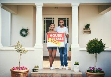 Shot of a young couple standing in front of their new home with a sold real estate sign